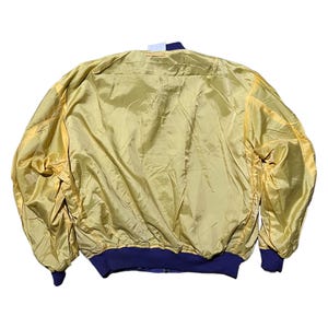 May include: A vintage gold bomber jacket with a shiny, satin-like finish. The jacket features navy blue ribbed cuffs and a matching collar. The back of the jacket is visible, showcasing its full shape and design.