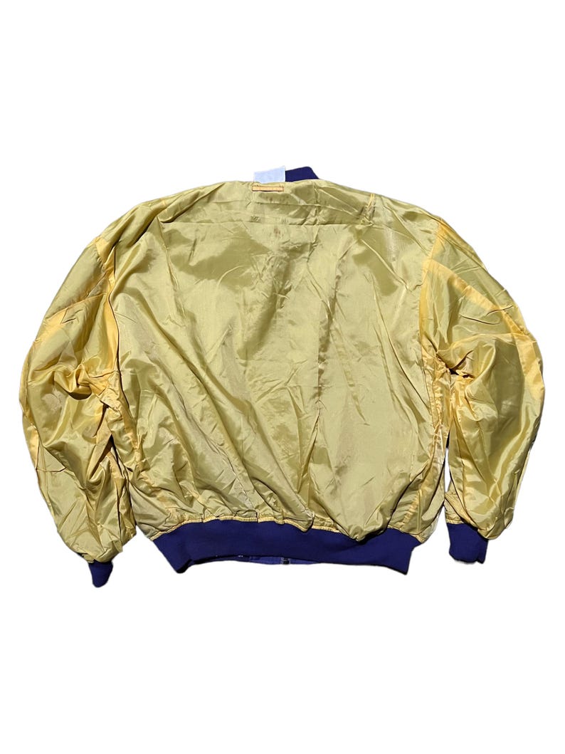 May include: A vintage gold bomber jacket with a shiny, satin-like finish. The jacket features navy blue ribbed cuffs and a matching collar. The back of the jacket is visible, showcasing its full shape and design.