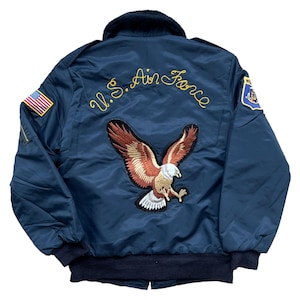 Può includere: Giacca bomber blu navy con colletto e polsini neri. Presenta un'ampia toppa con aquila ricamata sul retro, insieme alla scritta "U.S. Air Force" in oro. Include una toppa con la bandiera americana sulla manica sinistra.