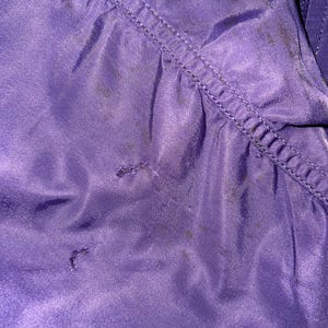May include: Close-up of a section of a purple fabric, possibly a jacket or coat. The fabric has a sheen and visible stitching along a seam. There are several dark spots on the material, and the texture appears smooth.