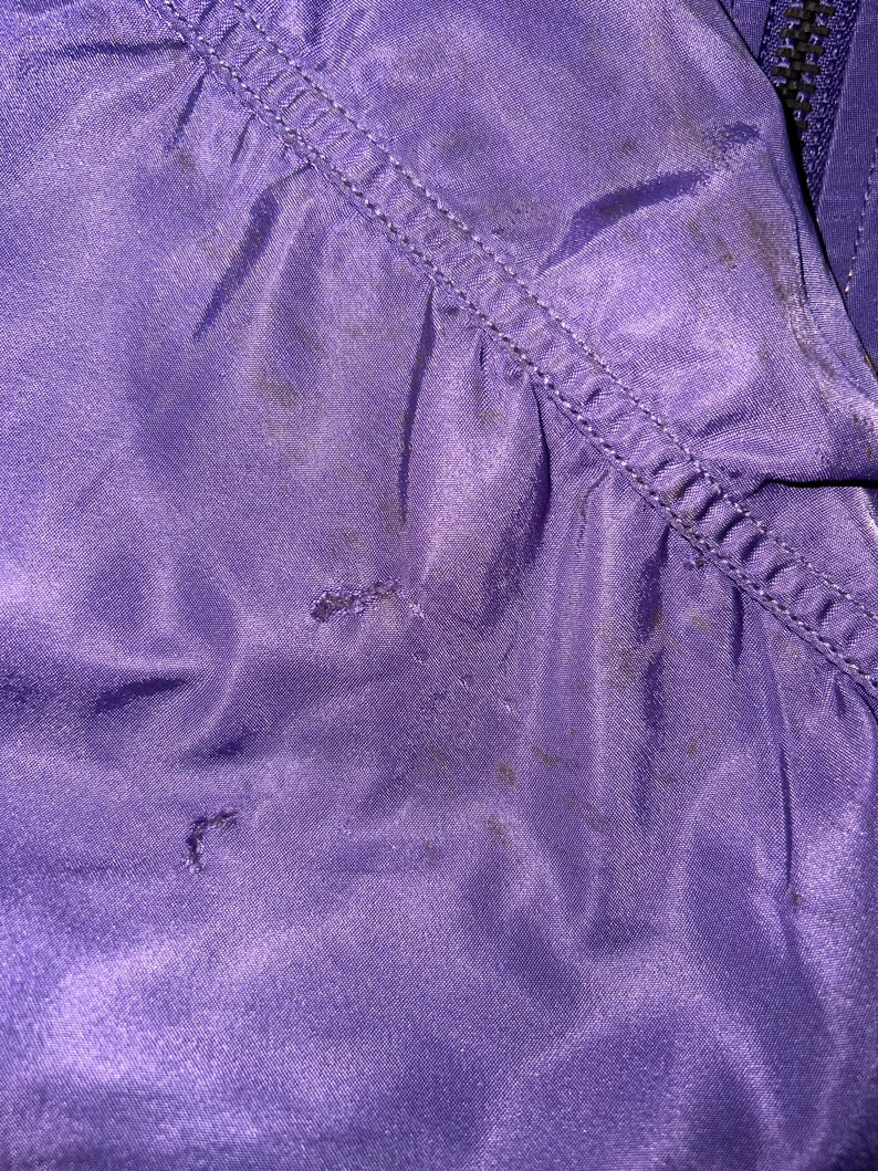 May include: Close-up of a section of a purple fabric, possibly a jacket or coat. The fabric has a sheen and visible stitching along a seam. There are several dark spots on the material, and the texture appears smooth.