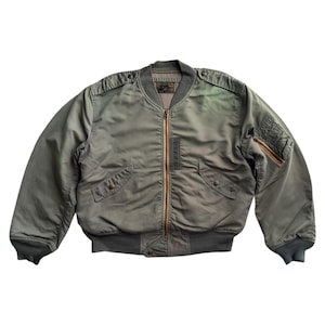 Może przedstawiać: Zielona kurtka bomberka w kolorze oliwkowym ze złotym zamkiem błyskawicznym, dwiema przednimi kieszeniami oraz prążkowanym kołnierzem i mankietami. Kurtka ma klasyczny, inspirowany wojskiem design.