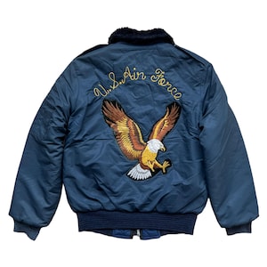 May include: Navy blue bomber jacket with a black faux fur collar. The back features an embroidered eagle with the words "U.S. Air Force" in gold thread. The jacket has a ribbed hem and cuffs.