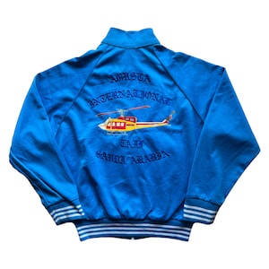 Può includere: Giacca blu vintage con collo alto e polsini a righe bianche. La parte posteriore presenta un design di elicottero ricamato con la scritta "Agusta International, TAJ, Saudi Arabia."