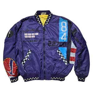 May include: Purple bomber jacket with a yellow lining and a black and white checkered collar. The jacket features patches with the number 84, the text "Big Beautiful Doll", and other graphics. The sleeves have zippered pockets and the text "Miss Miami".