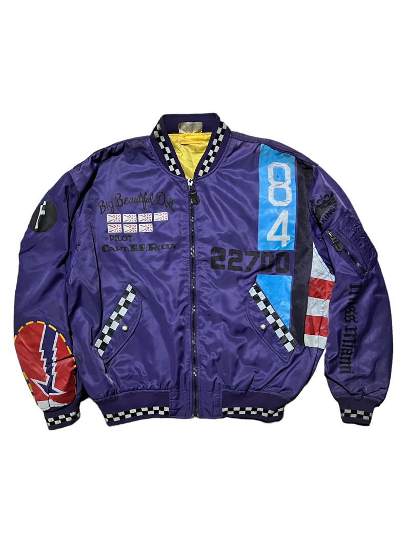 May include: Purple bomber jacket with a yellow lining and a black and white checkered collar. The jacket features patches with the number 84, the text "Big Beautiful Doll", and other graphics. The sleeves have zippered pockets and the text "Miss Miami".