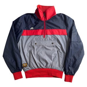 Peut inclure: Veste anorak vintage de couleur bleu marine, rouge et grise. La veste présente un col montant, une fermeture à demi-zip et une poche kangourou. Les manches et l'ourlet ont des poignets côtelés rouges. Un petit logo est visible sur la poitrine.