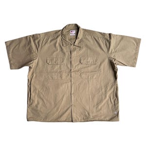 Può includere: Camicia beige a maniche corte con due tasche sul petto. La camicia ha un colletto ed è realizzata in un tessuto leggero. Il colore neutro è adatto per l'abbigliamento casual.