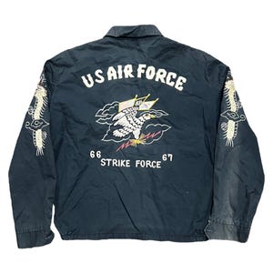 Może przedstawiać: Ciemnoniebieska kurtka US Air Force z haftowanym orłem i napisem. Tył zdobi biały orzeł z żółtymi akcentami, napis "US AIR FORCE" i "STRIKE FORCE 66 67". Rękawy mają białe hafty.
