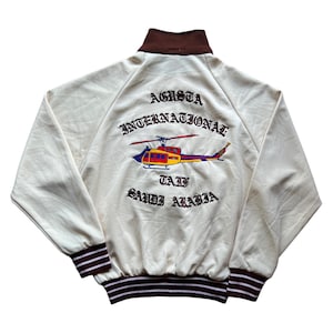 Può includere: Giacca bomber color crema con colletto marrone e polsini a righe. La parte posteriore presenta un design di elicottero ricamato con la scritta "Agusta International Taif Saudi Arabia" in stile vintage. La giacca ha una chiusura con zip completa.