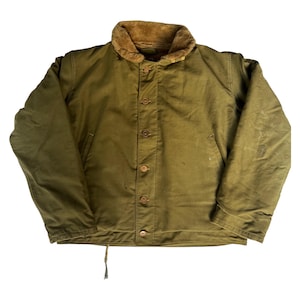 Può includere: Giacca vintage verde oliva con colletto in pelliccia marrone. La giacca ha una chiusura con bottoni sul davanti e due tasche anteriori. La giacca è di taglia media.