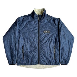 May include: A quilted navy blue jacket with a cream-colored lining and a high collar. The jacket features a full-length zipper, two side pockets, and the "Wild Things" logo on the chest. The jacket is designed with a diamond pattern.