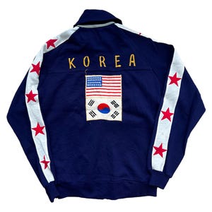 Może przedstawiać: Granatowa bluza z białymi paskami i czerwonymi gwiazdami na rękawach. Słowo "KOREA" jest wyhaftowane złotą nicią nad naszywką łączącą flagi amerykańską i południowokoreańską. Top w stylu vintage sportowym.
