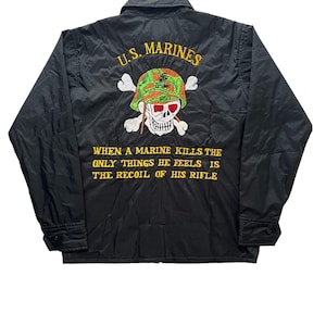 Może przedstawiać: Czarna nylonowa kurtka z haftowanym wzorem. Z tyłu znajduje się czaszka w hełmie z napisem "U.S. MARINES" i cytatem "WHEN A MARINE KILLS THE ONLY THINGS HE FEELS IS THE RECOIL OF HIS RIFLE."
