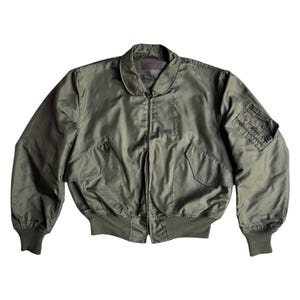 May include: An olive green bomber jacket with a ribbed collar, cuffs, and hem. The jacket has two front pockets, a zip closure, and a small pocket on the left sleeve. The jacket is made of a smooth material, possibly nylon.