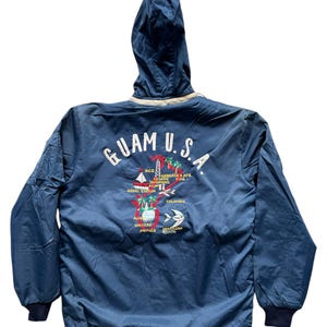 May include: A navy blue hooded jacket with the text "GUAM U.S." embroidered on the back, along with various illustrations and place names. The jacket has a white trim around the hood and navy blue cuffs.