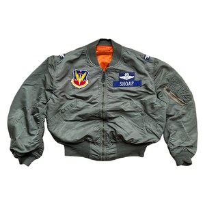 Puede incluir: Chaqueta bomber verde oliva con forro naranja. Cuenta con parches con el texto "SHOAF" y un emblema militar. Incluye un bolsillo con cremallera en la manga izquierda y puños y cintura de canalé. Una chaqueta clásica de estilo militar.