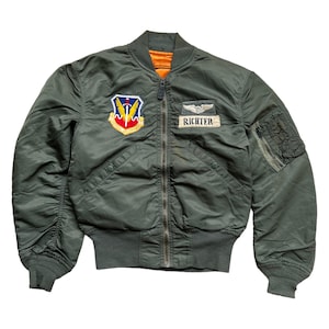 Puede incluir: Chaqueta bomber verde oliva con cuello, puños y dobladillo de canalé. Cuenta con un parche con un escudo y alas, y una etiqueta con el nombre que dice "RICHTER". La chaqueta tiene cierre de cremallera y un bolsillo en la manga izquierda.