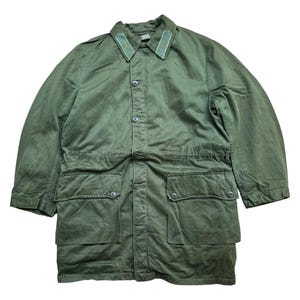 Puede incluir: Una chaqueta larga de manga larga de estilo militar de color verde con cierre de botones y dos bolsillos grandes. La chaqueta tiene una cintura con cordón y un cuello con ribete verde y dorado.