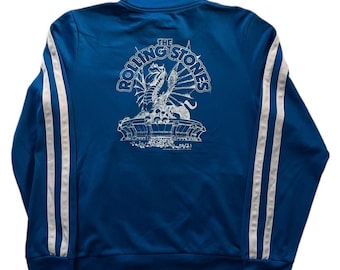 Puede incluir: Chaqueta deportiva azul con rayas blancas en las mangas. La parte trasera presenta un gráfico blanco de un dragón y las palabras "The Rolling Stones". La chaqueta tiene cremallera completa y cuello alzado.