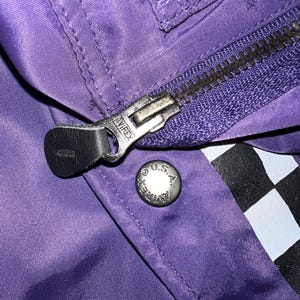 May include: Close-up of a purple jacket with a silver zipper and a snap button. The zipper pull has the brand name "AVIREX" embossed on it. The snap button is silver with the text "AVIREX U.S.A." around the edge. A black and white checkered pattern is visible.