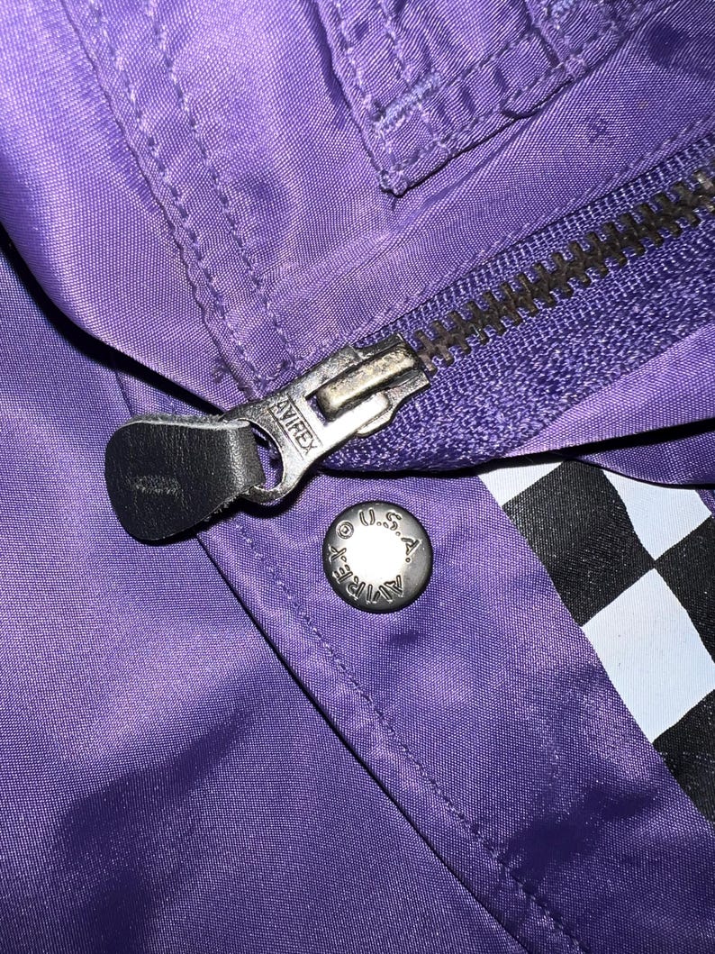 May include: Close-up of a purple jacket with a silver zipper and a snap button. The zipper pull has the brand name "AVIREX" embossed on it. The snap button is silver with the text "AVIREX U.S.A." around the edge. A black and white checkered pattern is visible.
