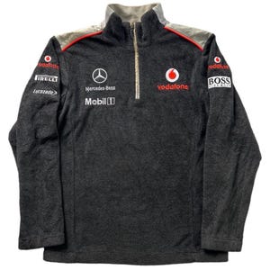 May include: A dark gray fleece pullover with a half-zip collar. The pullover features the Vodafone logo, Mercedes-Benz logo, and sponsor logos like Pirelli and BOSS. The shoulders have a gray and red accent.