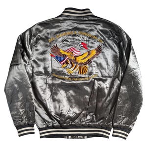 Może przedstawiać: Srebrna satynowa kurtka bomberka z czarno-białym kołnierzem, mankietami i obszyciem w paski. Z tyłu haftowany orzeł z amerykańską flagą i napisem "USA RICHMOND K. TURNER (06-20)" oraz "MULTINATIONAL PEACEKEEPING FORCE".