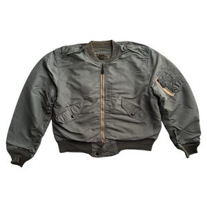 Może przedstawiać: Szałwiowa kurtka bomberka z brązowym zamkiem błyskawicznym i wieloma kieszeniami. Kurtka ma prążkowany kołnierz, mankiety i pas. Klasyczny styl.