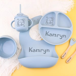 May include: A blue silicone baby feeding set with a plate, bowl, cup, bib, and two spoons. The set features a space-themed design with a planet and stars. The items are personalized with the name "Kamryn".