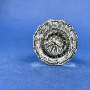 May include: A clear glass door knob featuring a multi-faceted, starburst-like pattern radiating from the center. The knob has a circular, scalloped edge design with numerous reflective surfaces that catch light, creating a complex geometric appearance.