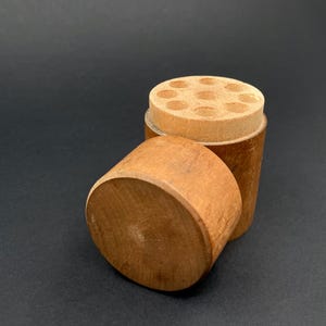 May include: A small, cylindrical wooden container with a removable lid. The lid has multiple small holes. The container is made of light brown wood and sits on a dark background. The lid is off to the side.