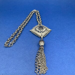 May include: Silver-toned necklace with a diamond-shaped pendant and tassel. The pendant features a detailed design with a dark center. The chain is a classic link style, and the background is a solid blue.