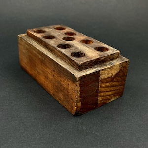May include: A rectangular wooden desk organizer with a dark brown finish. The organizer has a stepped design with a top section featuring ten evenly spaced circular holes. The wood grain is visible, adding texture to the piece.