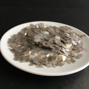 May include: A white ceramic plate filled with shimmering, gray mica flakes. The flakes have a layered, irregular shape, and reflect light, creating a sparkling effect. The background is a solid black.