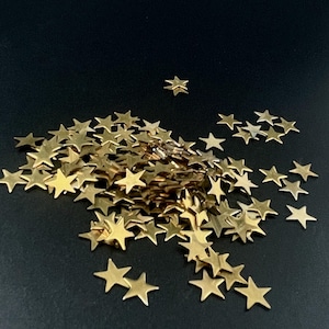 May include: A collection of small, flat, five-pointed star-shaped sequins in a metallic gold finish. The stars are scattered in a loose pile, with some individual pieces separated from the main cluster, all featuring a smooth, reflective surface.