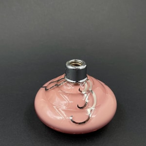 May include: A pink glass perfume bottle with a silver metal top. The bottle has a rounded shape and is decorated with silver swirls. The top is black and silver. The bottle is empty.