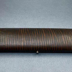 May include: A rectangular, wooden box with a dark brown finish and a striped pattern. The box has a small, silver-colored clasp. The box is likely used for storing jewelry or other small items.