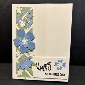 May include: A white Mother's Day card with a floral design. The card features blue and white flowers, green leaves, and the words "Happy Mother's Day" in black script. The card has a decorative border.