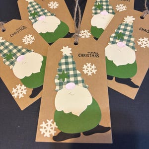 May include: A collection of six brown paper gift tags with a gnome design. Each tag features a green and white plaid hat, a white beard, and a green body. The tags are adorned with snowflake cutouts and the words "Merry Christmas".