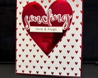 May include: A red heart-shaped card with a white background and a red heart pattern. The card says "sending love & hugs" in red lettering.