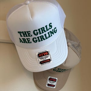 May include: White trucker hat with the text "THE GIRLS ARE GIRLING" in green. The hat has a white mesh back and a curved brim. A second tan hat with the same design is visible below.