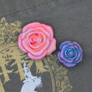 May include: A close-up of a gray tote bag with a gold-colored emblem featuring a mouse wearing a wizard hat. Two rose-shaped pins, one pink and one purple, are placed on the bag. The background is a blue and white tie-dye pattern.