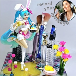 Puede incluir: Una obra de arte de resina colorida con una figura de un personaje de anime de cabello azul que lleva un disfraz de conejito blanco y sostiene una cesta de huevos de Pascua. La figura está rodeada de pinceles de maquillaje, botellas de perfume y una pequeña figura de pollito amarillo. El texto "record your message" está escrito en letras blancas sobre fondo negro.