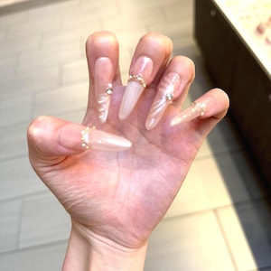 May include: A hand with long, light pink acrylic nails. Each nail is adorned with gold rings and small, sparkling embellishments. The nails have a glossy finish and a natural, elegant appearance.