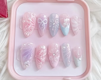 Könnte beinhalten: Ein Set mit zehn falschen Nägeln in einem rosa Plastiketui. Die Nägel sind mit weißen und violetten Blumenmustern und Strasssteinen verziert.