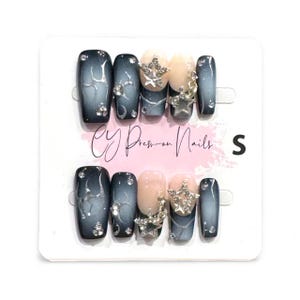 May include: A set of press-on nails with a gradient design in shades of blue and gray, adorned with silver star and rhinestone embellishments. Some nails are a nude color. The text "CY Press-on Nails" is visible.