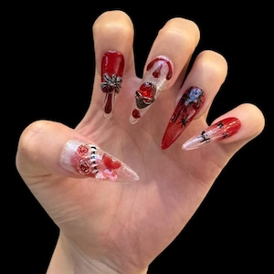 May include: A hand displaying long, pointed, artificial nails with a dark, gothic theme. The nails feature a variety of designs, including red and black accents, spider and heart embellishments, and floral details. The nails are set against a black background.