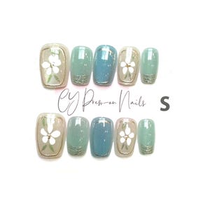 Puede incluir: Un conjunto de uñas postizas a presión en varios tonos de azul y beige, con un diseño floral. Algunas uñas tienen un acento de purpurina en la base. El texto "CY Press-on Nails" es visible, junto con la letra "S".