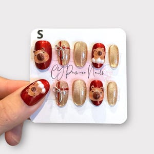 May include: A set of press-on nails in various sizes. The nails feature a festive design with red, gold, and brown colours. Some nails have a 3D bear design, while others have a bow or glitter. The text "CY Press-on Nails" is visible.